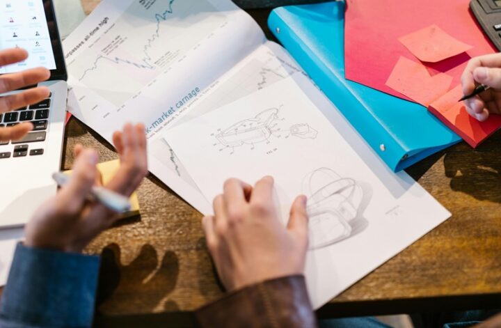Business team discussing funding strategy with charts, product sketches, and documents on a desk.