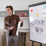 A startup founder presents growth data beside a whiteboard labeled “STARTUP,” discussing funding goals and business milestones.