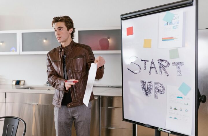A startup founder presents growth data beside a whiteboard labeled “STARTUP,” discussing funding goals and business milestones.