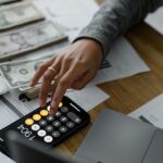 Hand using a calculator on a smartphone while reviewing financial documents and cash, representing budgeting and funding goal planning.