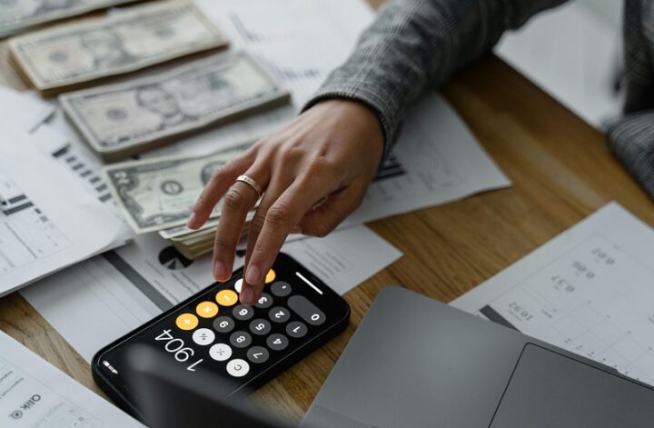 Hand using a calculator on a smartphone while reviewing financial documents and cash, representing budgeting and funding goal planning.